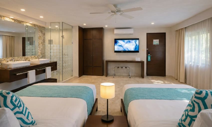 Habitación doble de hotel con dos camas, detalles turquesas, televisión y baño integrado.