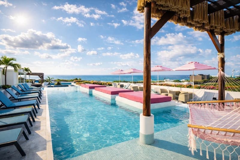 Alberca infinita en la azotea con camastros rosas, hamaca y vista panorámica al mar Caribe.